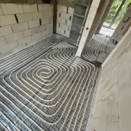 Białe rury ogrzewania podłogowego ułożone spiralnie na srebrnej folii izolacyjnej z nadrukiem siatki, widoczne surowe ściany z betonu komórkowego i okno w tle.