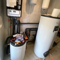 SAN-BUD CONSTRUCTIONS SPÓŁKA Z OGRANICZONĄ ODPOWIEDZIALNOŚCIĄ - Biały piec gazowy Vaillant z podłączonymi rurami, na jego górze rozłożone narzędzia hydrauliczne i pudełko złączek, obok drugi, większy zbiornik, na ścianie wiszący moduł sterujący