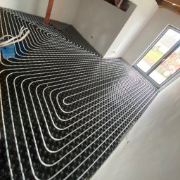 SAN-BUD CONSTRUCTIONS SPÓŁKA Z OGRANICZONĄ ODPOWIEDZIALNOŚCIĄ - Białe rurki ułożone spiralnie na czarnej macie w trakcie montażu ogrzewania podłogowego w surowym pomieszczeniu z oknem.