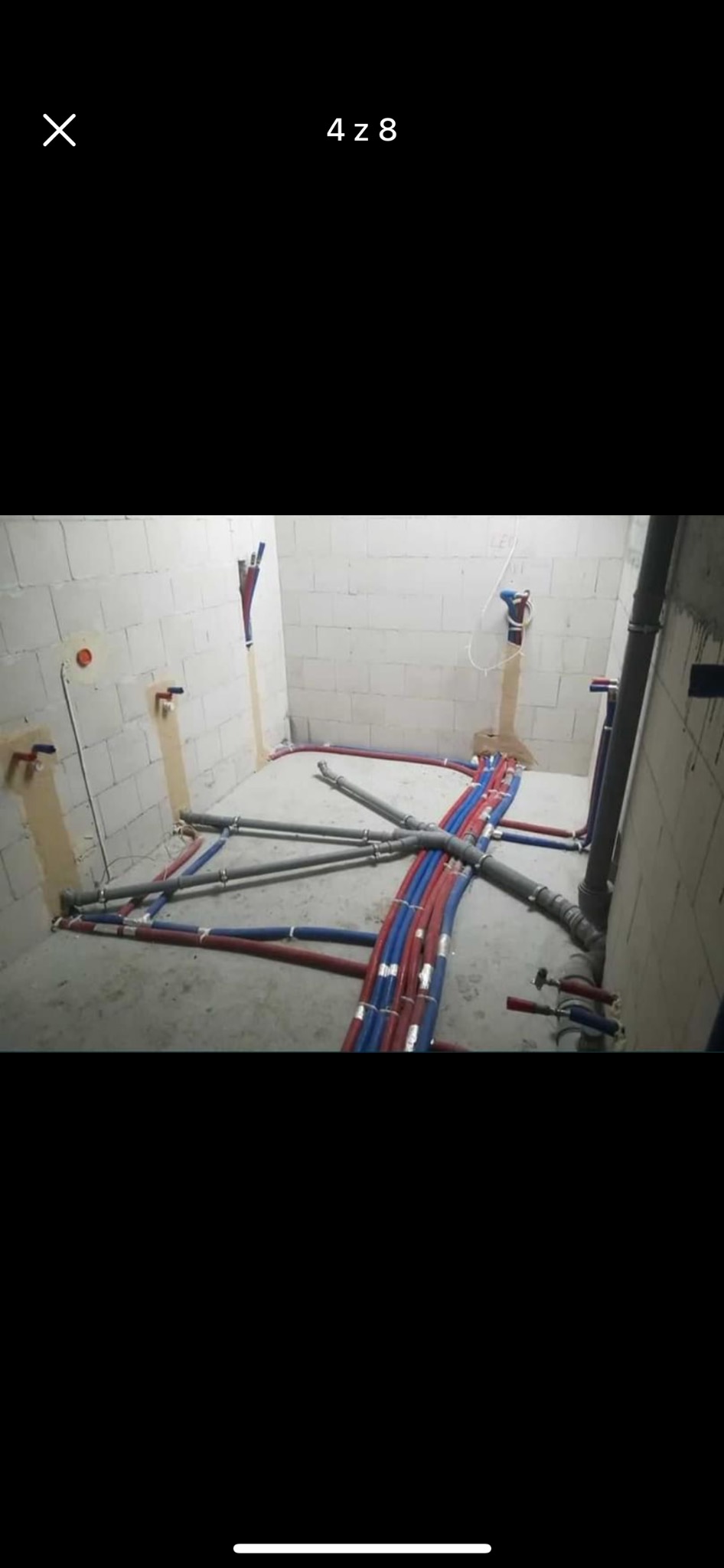 Surowe wnętrze z widoczną siecią rur grzewczych i wodociągowych w trakcie instalacji, czerwone i niebieskie rury ułożone na posadzce, ściany z betonu komórkowego.