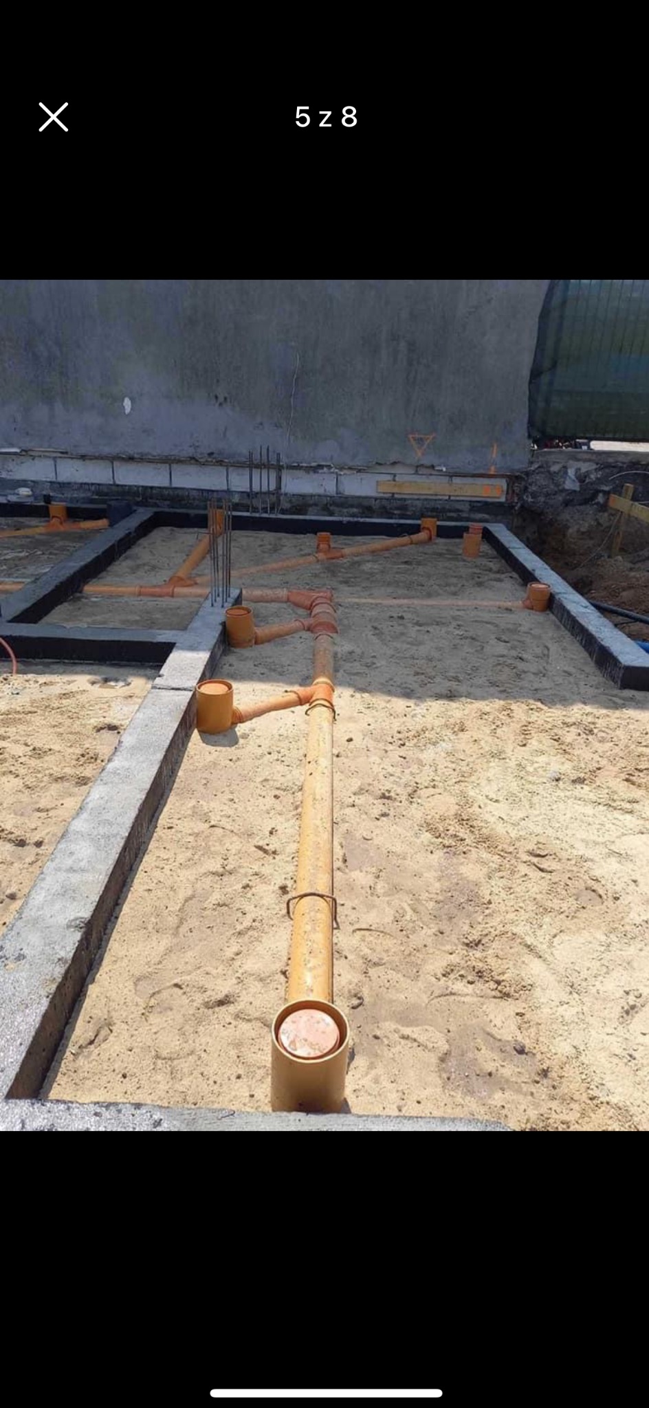 Pomarańczowe rury kanalizacyjne ułożone w piasku w fundamencie budynku, widoczne zbrojenia i betonowe ławy.