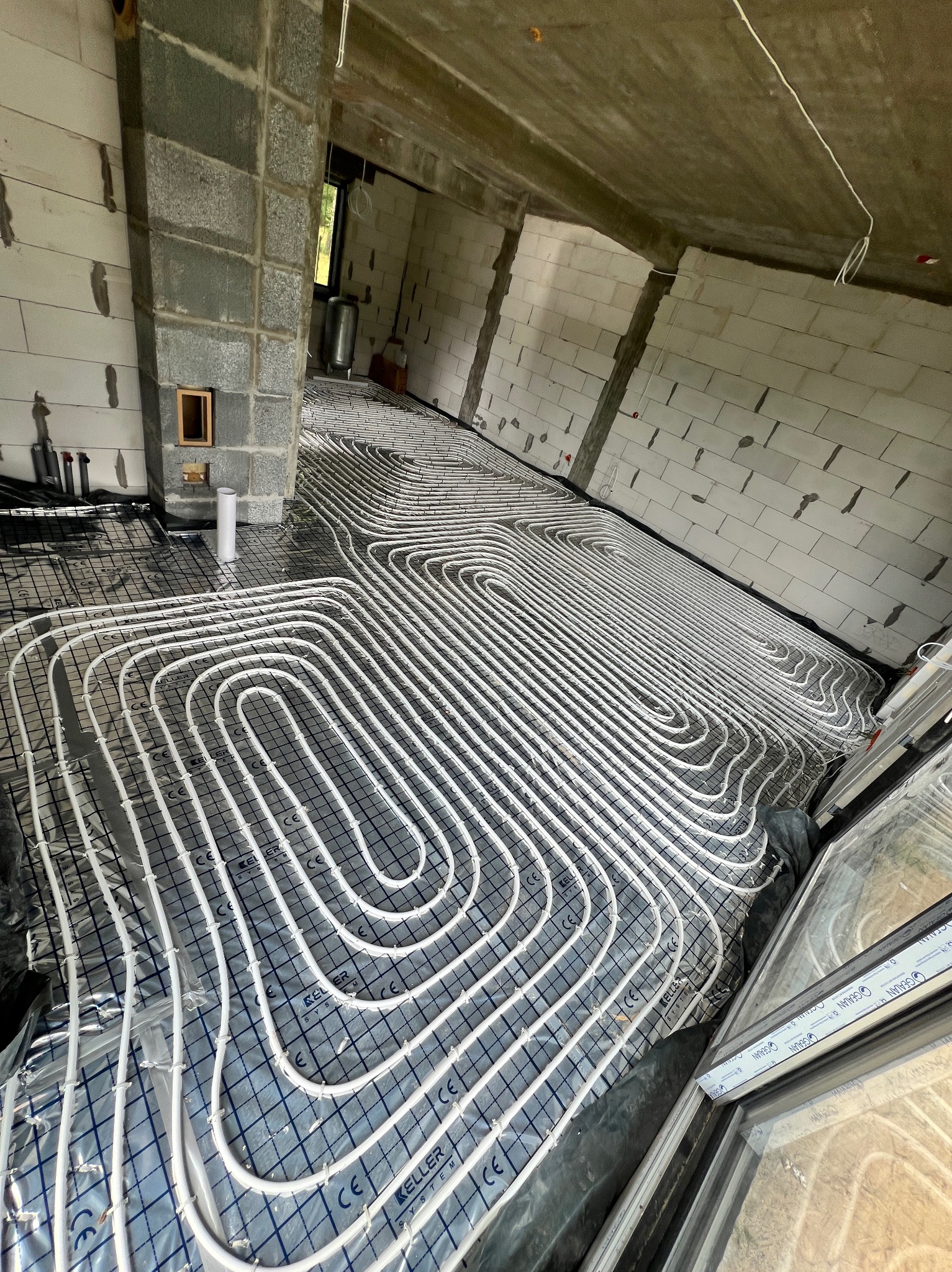 Rozłożone białe rurki ogrzewania podłogowego na folii z siatką w surowym wnętrzu budynku z bloczków, widoczne okna i pion kanalizacyjny.