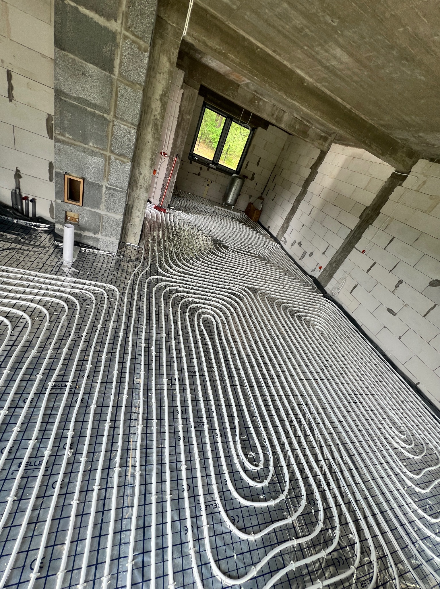 Białe rury ogrzewania podłogowego ułożone na siatce w surowym wnętrzu budynku z betonowymi ścianami i oknem w tle.