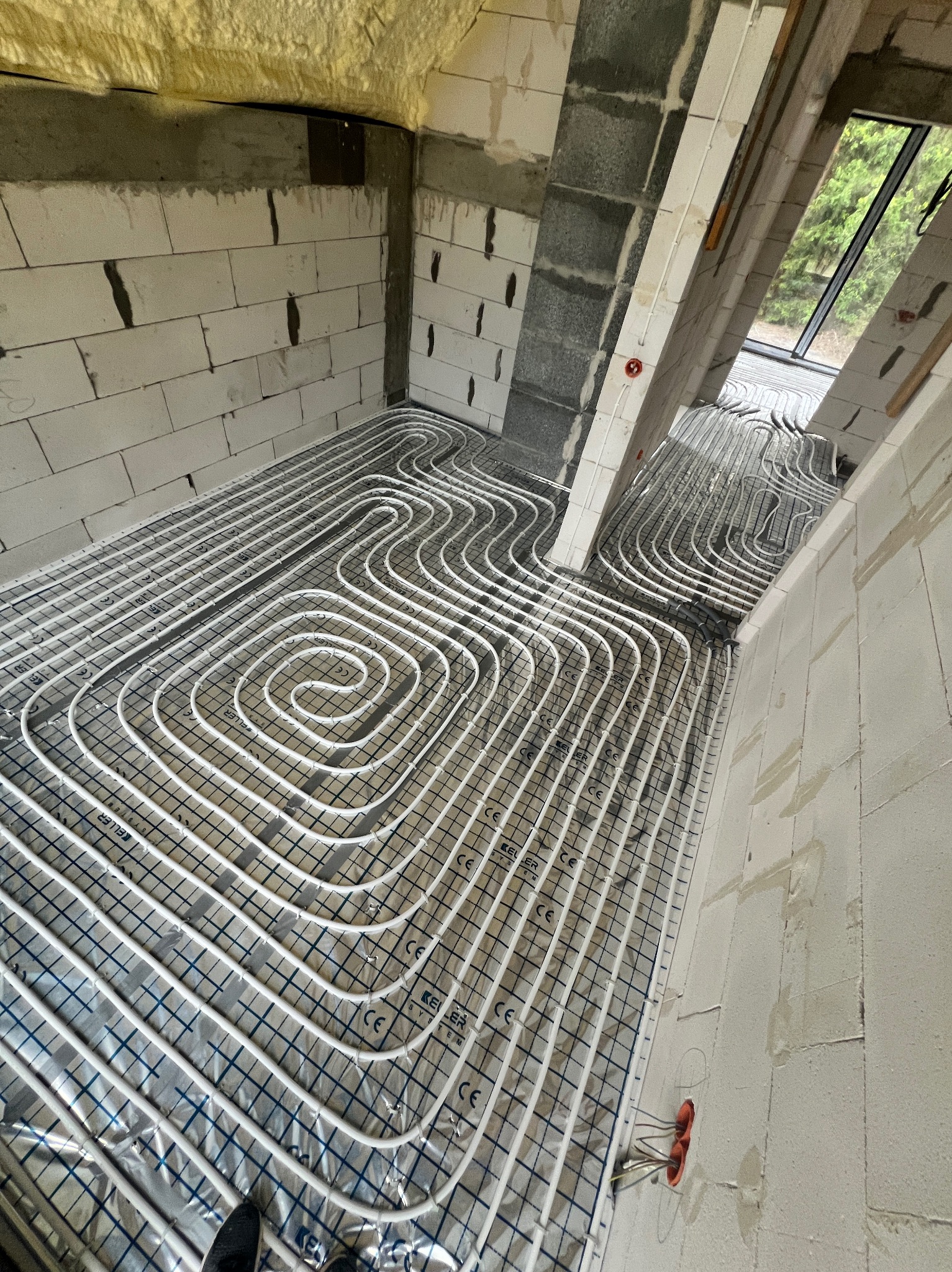 Białe rury ogrzewania podłogowego ułożone spiralnie na srebrnej folii izolacyjnej z nadrukiem siatki, widoczne surowe ściany z betonu komórkowego i okno w tle.