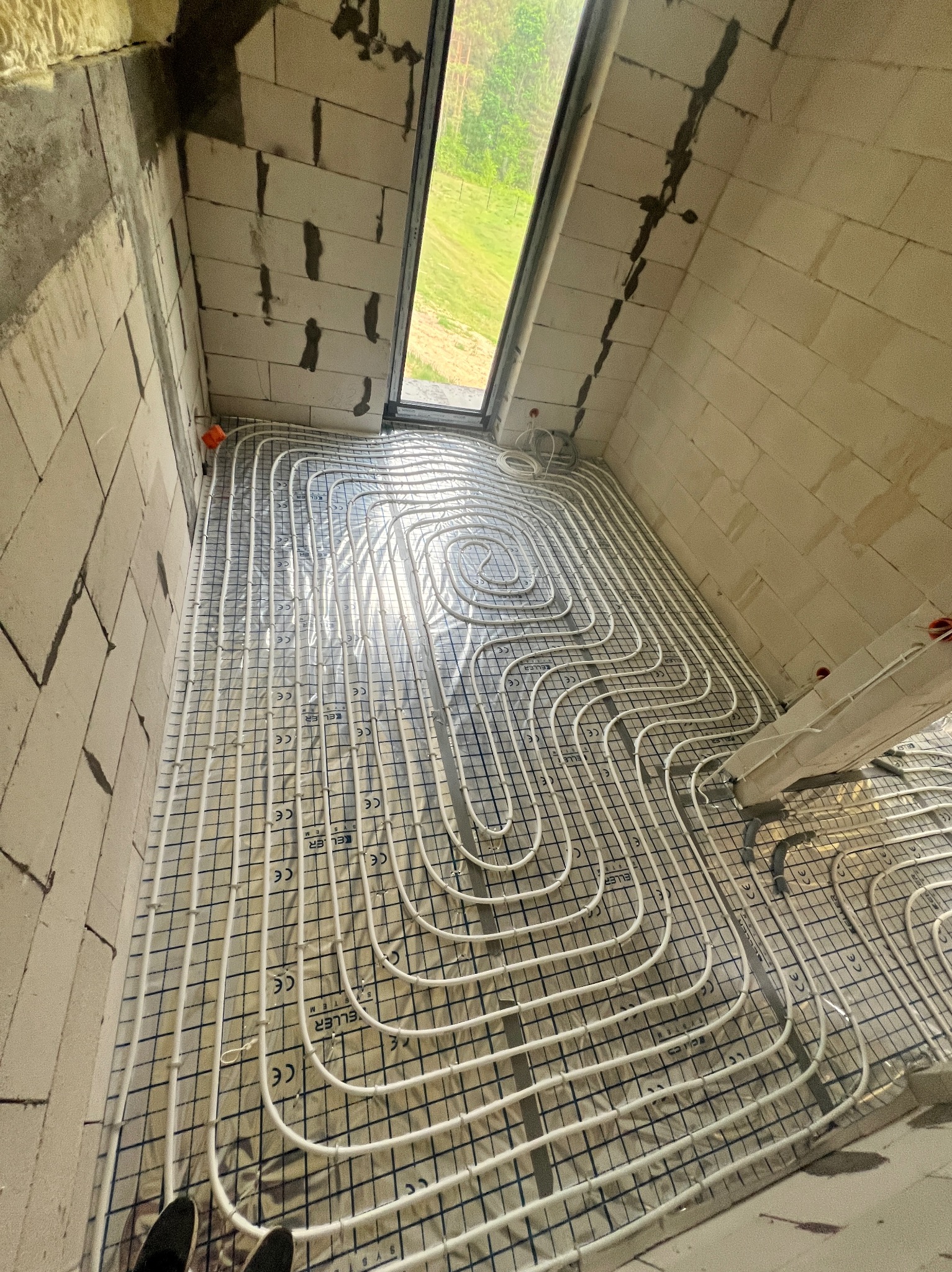 Instalacja ogrzewania podłogowego w trakcie realizacji w Płońsku, widok z góry na rozłożone rury na izolacji z siatką, surowy stan ścian z betonu komórkowego, widoczne fragmenty obuwia instalatora.