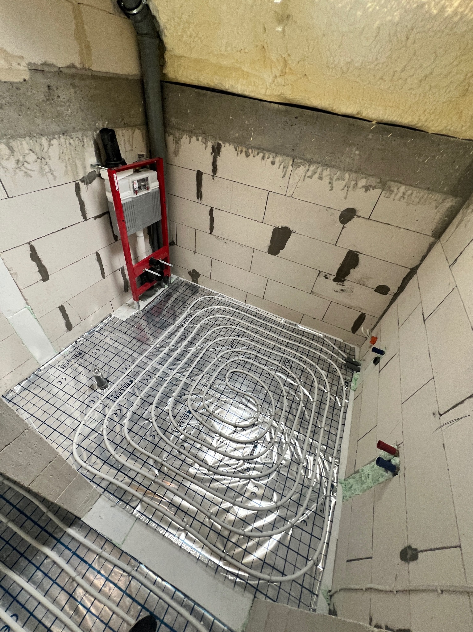 Białe rury ogrzewania podłogowego ułożone spiralnie na srebrnej folii z niebieską siatką w surowym pomieszczeniu z białych bloczków, widoczna rura odpływowa i stelaż podtynkowy WC.