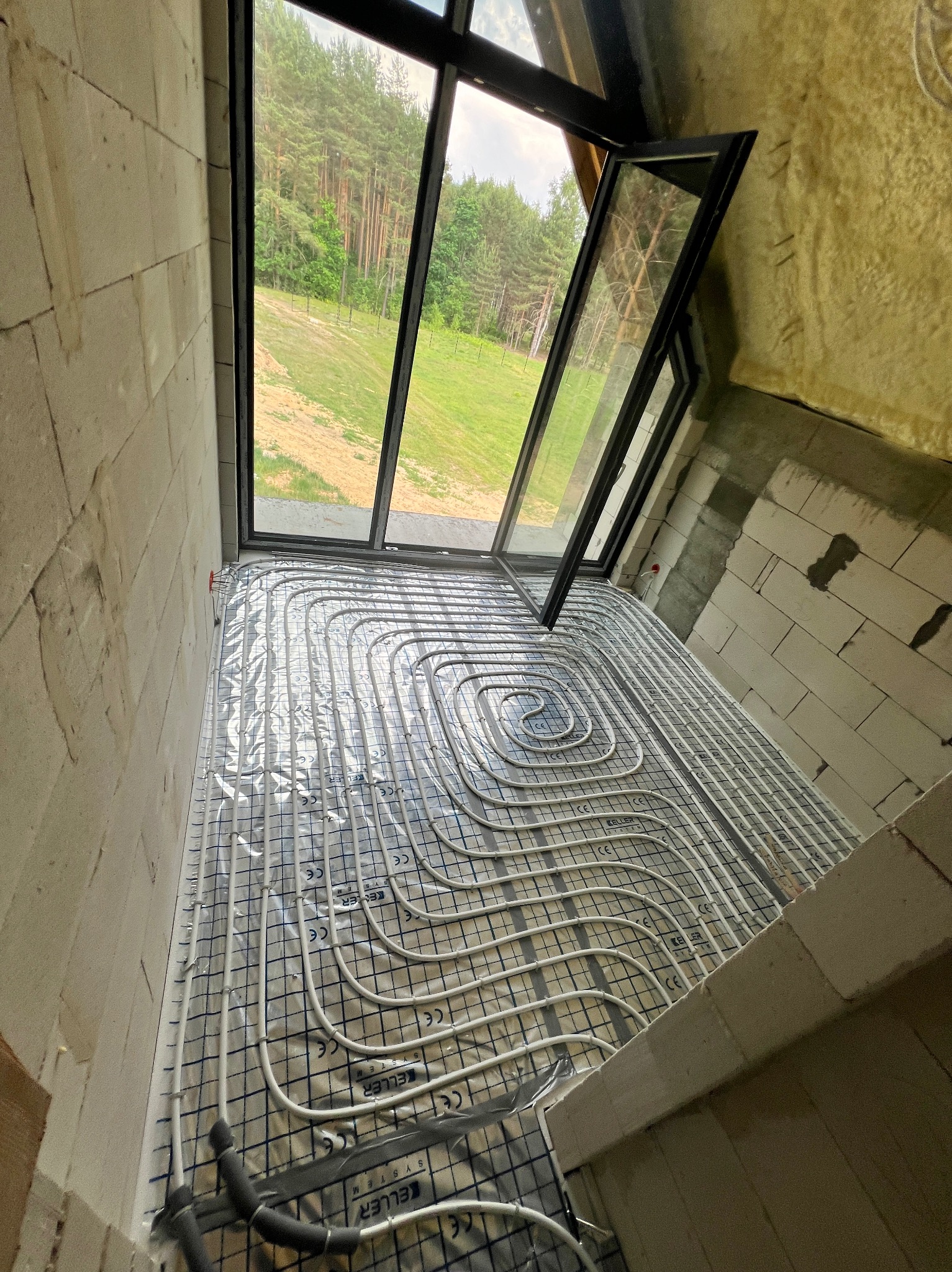 Montaż rur grzewczych w systemie ogrzewania podłogowego, widok z góry na rozłożone białe rury na folii z nadrukiem 'KELLER SYSTEM', surowe ściany z bloczków, otwarte okno dachowe z widokiem na las.