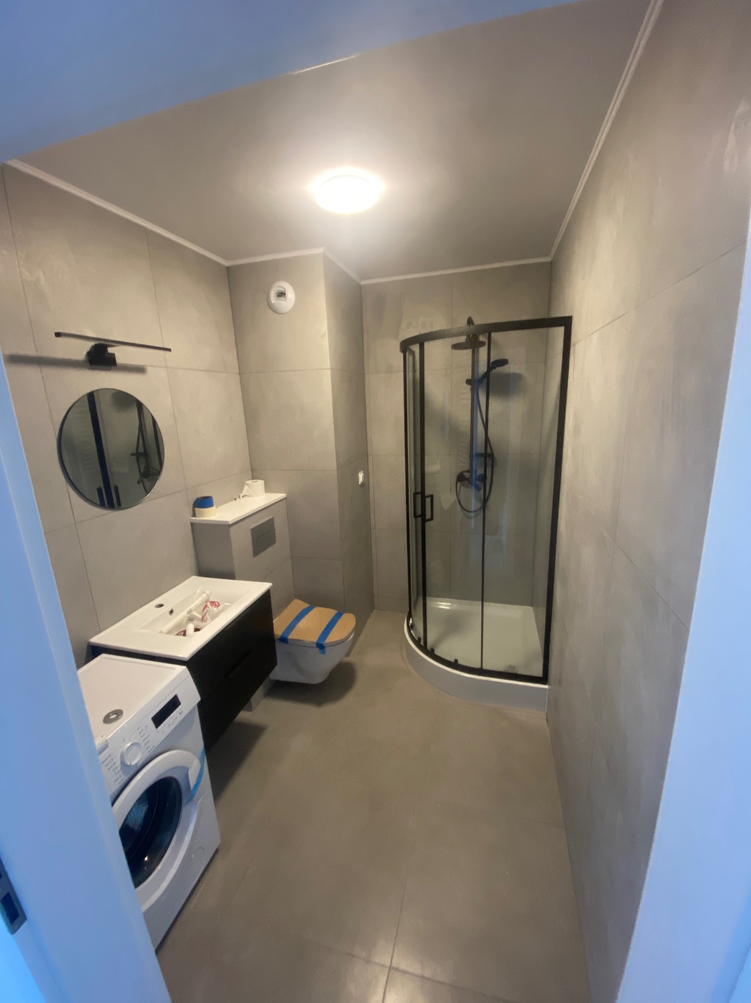 Wykończona łazienka w szarościach z kabiną prysznicową z czarnymi ramami, umywalką z szafką, toaletą zabezpieczoną taśmą i pralką z boku.