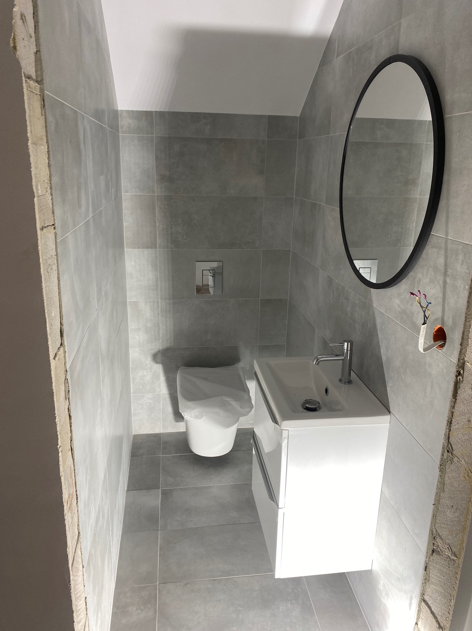 Wąska łazienka w trakcie remontu, szare płytki na ścianach i podłodze, biała umywalka z szafką i bateria, owalne lustro w czarnej ramie, toaleta w folii ochronnej, wystające kable ze ściany.