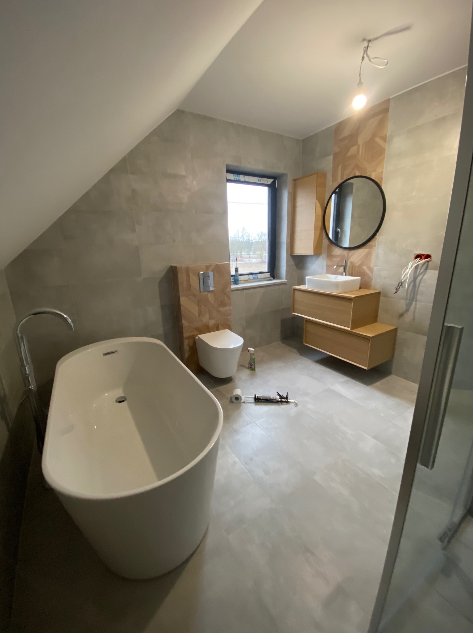 Nowoczesna łazienka na poddaszu ze skośnym sufitem, z wolnostojącą białą wanną, wiszącą toaletą z drewnianą obudową, umywalką na drewnianej szafce i okrągłym lustrem, na podłodze leżą narzędzia...