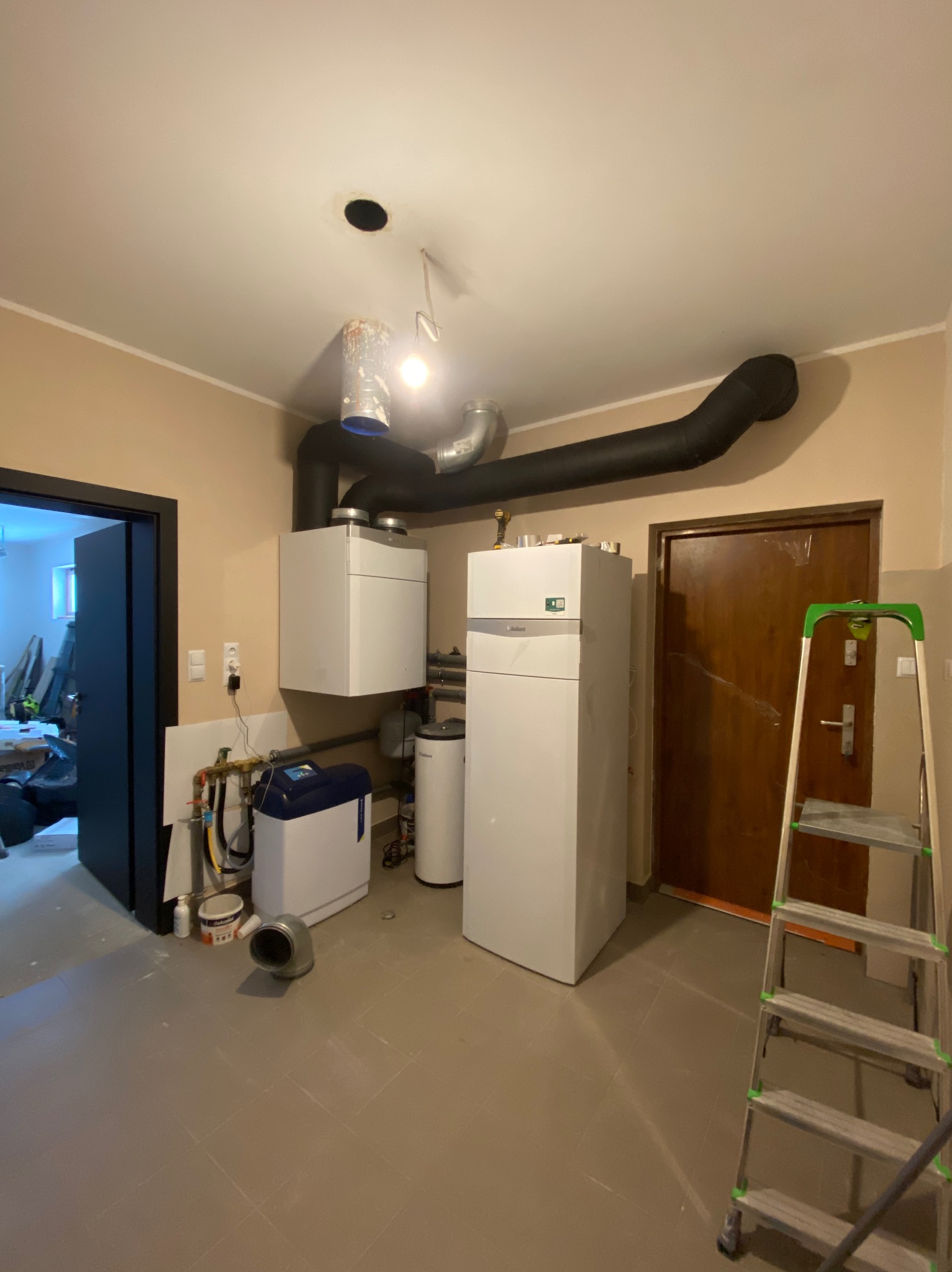 Pomieszczenie z zainstalowanym piecem gazowym wiszącym i stojącym, rurami odprowadzającymi spaliny, obok stoi drabina, widoczne elementy instalacji hydraulicznej.