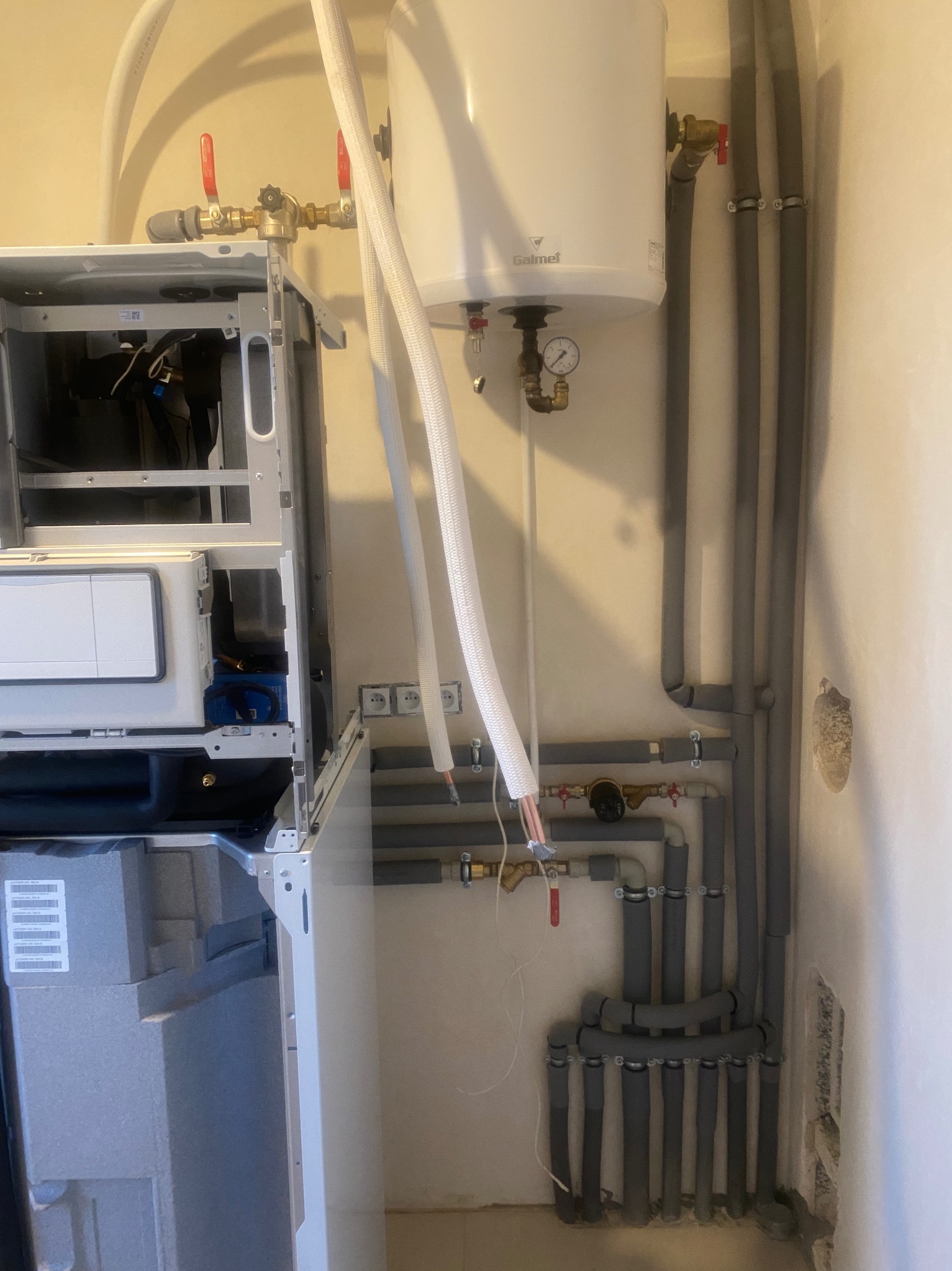 Wnętrze kotłowni w trakcie montażu, widoczny otwarty piec gazowy, bojler Galmet z podłączonymi rurami w izolacji termicznej i zaworami, instalacja elektryczna w ścianie.