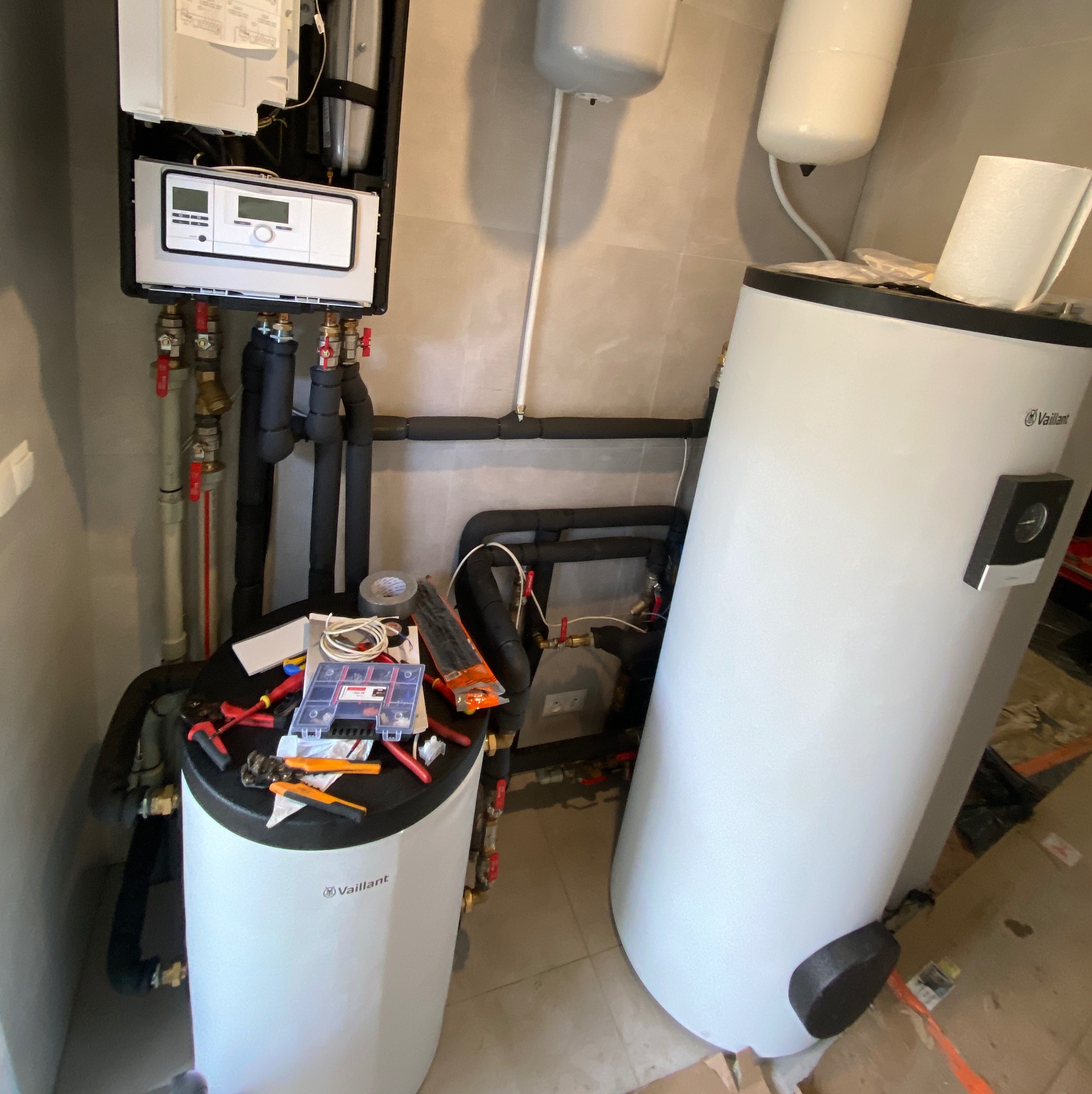 Biały piec gazowy Vaillant z podłączonymi rurami, na jego górze rozłożone narzędzia hydrauliczne i pudełko złączek, obok drugi, większy zbiornik, na ścianie wiszący moduł sterujący