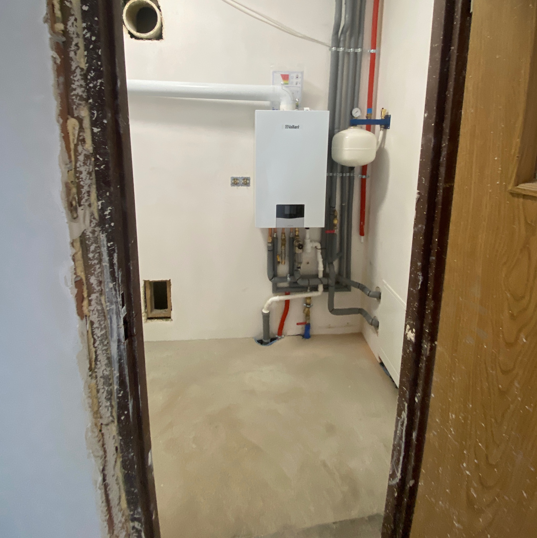 Nowy piec gazowy Vaillant zamontowany na ścianie, z podłączonymi rurami i zbiornikiem wyrównawczym, widoczne niedokończone wykończenie ościeżnicy drzwi i surowa wylewka podłogowa.
