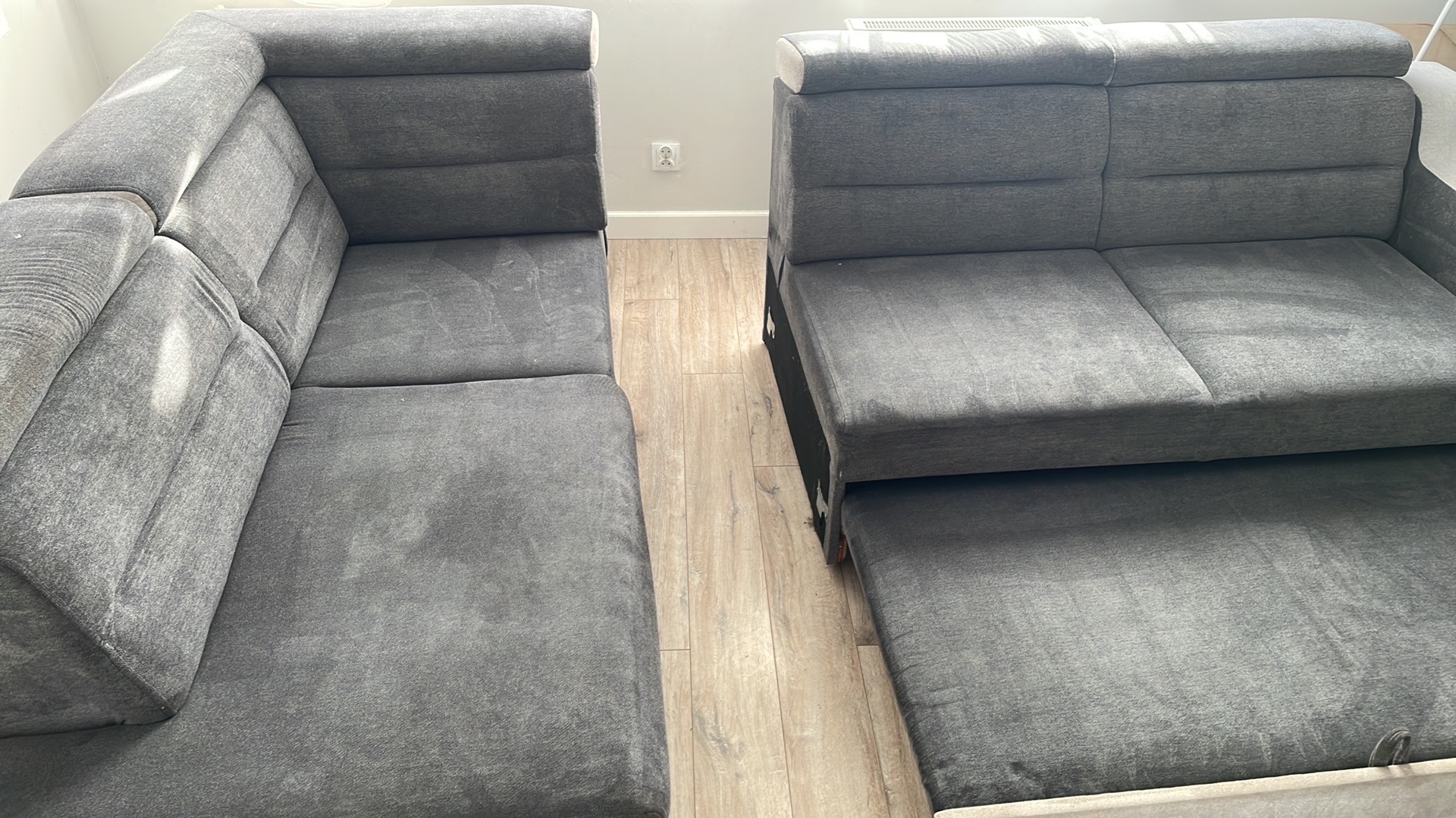 Szara, modułowa sofa narożna z wysuwanym elementem do spania, widoczna na drewnianej podłodze. Sofa po lewej stronie ustawiona w rogu pokoju.