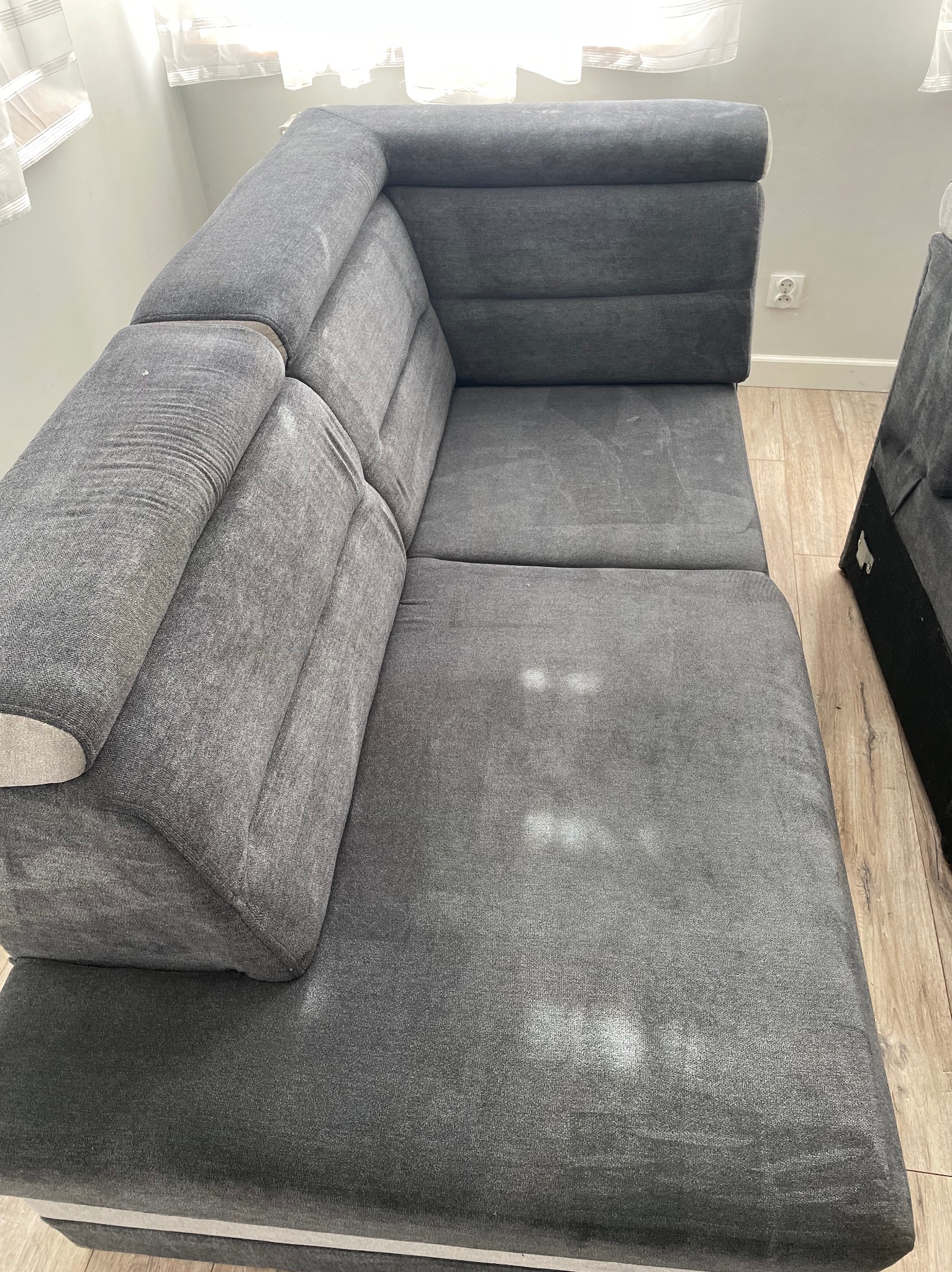 Szary narożnik z widocznymi plamami na tapicerce, stojący w rogu pokoju z drewnianą podłogą.