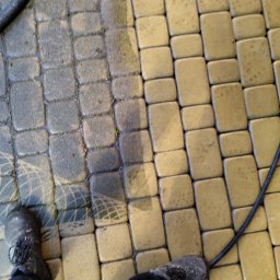 Mateusz Buczek - Częściowo umyta kostka brukowa w odcieniach żółci i szarości, widoczne różnice w czystości, w kadrze fragment węża ciśnieniowego i buty osoby wykonującej czyszczenie.