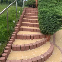 Schody zewnętrzne wyłożone kostką brukową w dwóch kolorach, z metalową barierką i ozdobnymi kulami świetlnymi w trawie, prowadzące do budynku z zielenią po bokach.