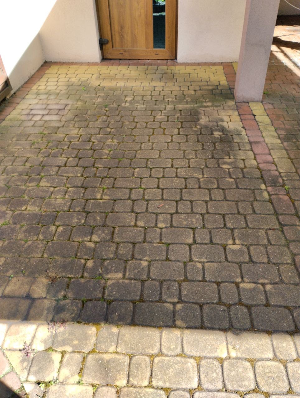 Zabrudzona kostka brukowa przed wejściem do domu, widoczne ślady wilgoci i porostów, część kostki jest wyraźnie jaśniejsza, co sugeruje niedawne czyszczenie.