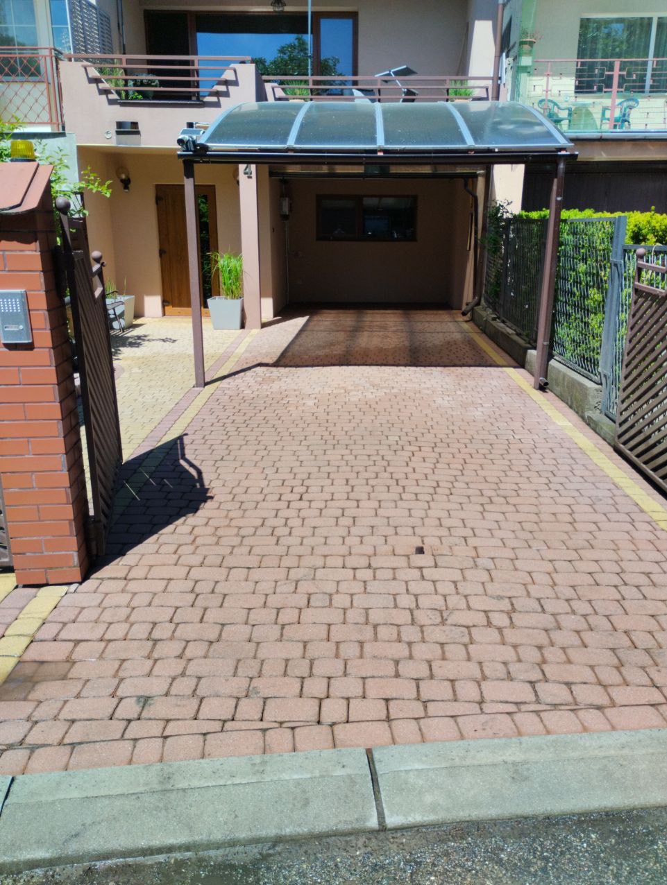 Czerwona kostka brukowa na podjeździe do garażu z zadaszeniem, widoczne ślady po niedawnym czyszczeniu, ciemniejszy obszar w miejscu parkowania samochodu.