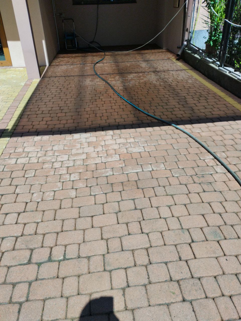 Częściowo umyta kostka brukowa na podjeździe z widocznym wężem ogrodowym i różnicą w kolorze między czystą a brudną powierzchnią.