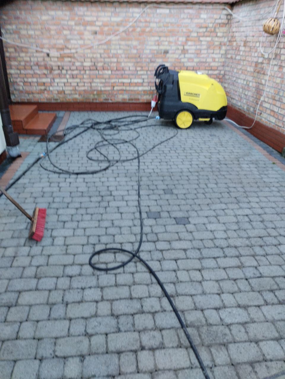 Żółto-czarna myjka ciśnieniowa Karcher na szarej kostce brukowej, obok leży czerwona szczotka, a w tle widać ceglany mur.
