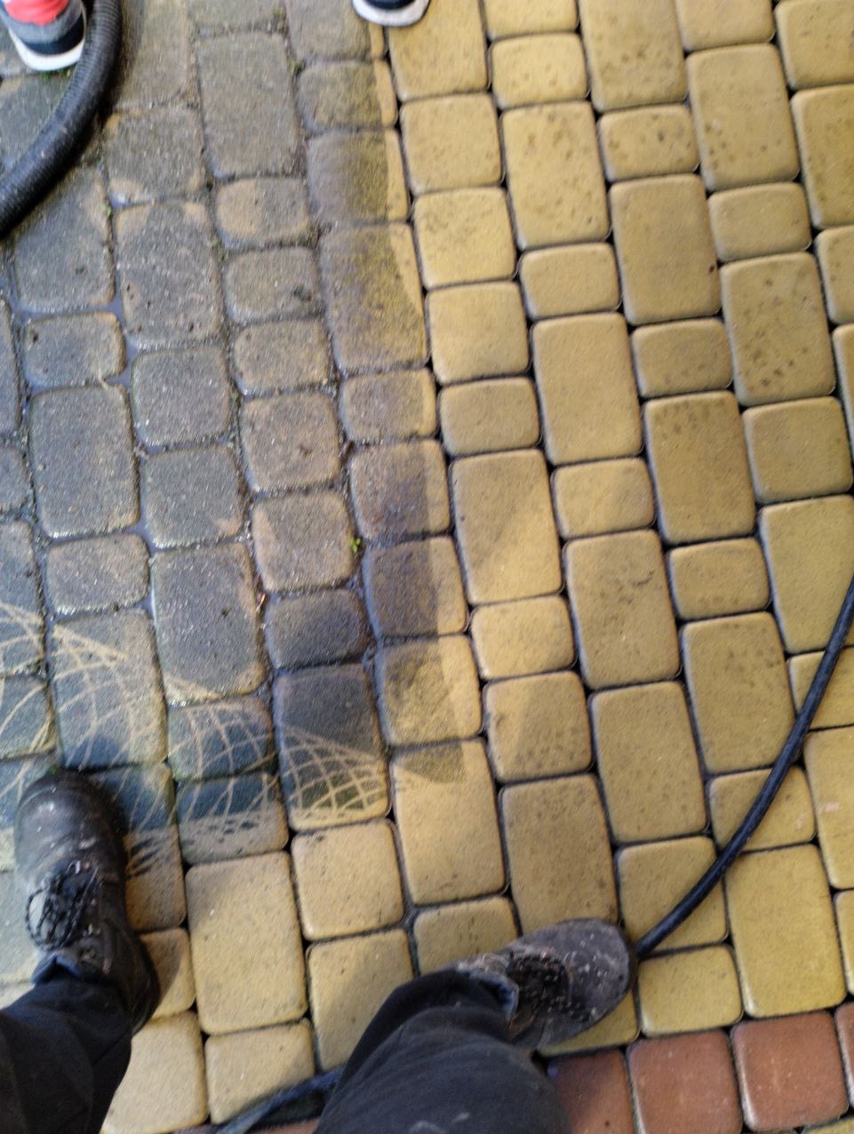 Częściowo umyta kostka brukowa w odcieniach żółci i szarości, widoczne różnice w czystości, w kadrze fragment węża ciśnieniowego i buty osoby wykonującej czyszczenie.
