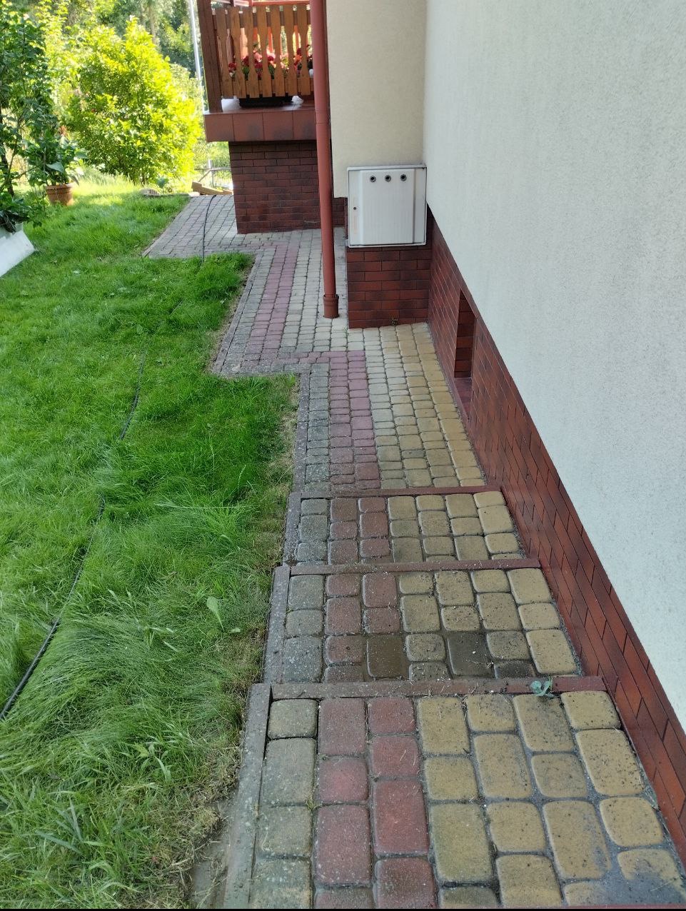 Częściowo umyta, kolorowa kostka brukowa prowadząca do budynku z białą elewacją, widoczne różnice w kolorze kostki przed i po czyszczeniu.
