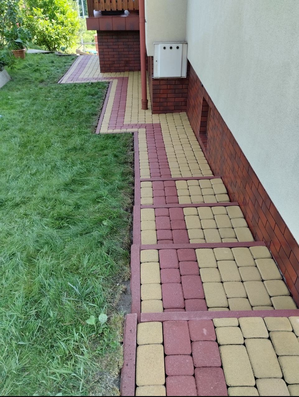 Ułożona kostka brukowa w dwóch kolorach (czerwonym i beżowym) tworząca ścieżkę i schody przy budynku z ceglaną podmurówką i zielonym trawnikiem.