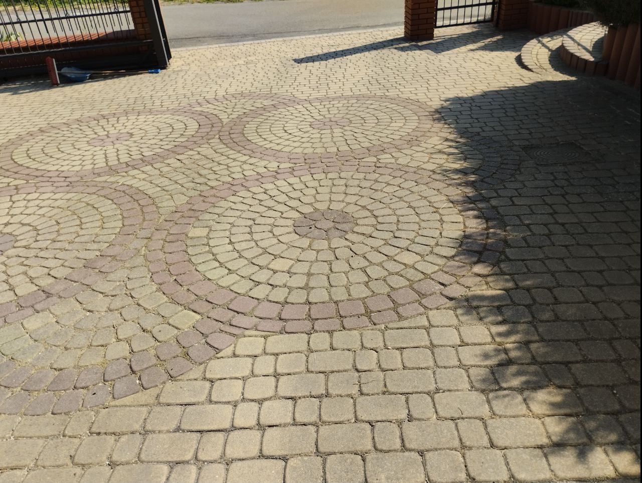 Ułożona kostka brukowa w kształcie okręgów, z widocznymi śladami cienia drzewa, prowadząca do bramy wjazdowej.