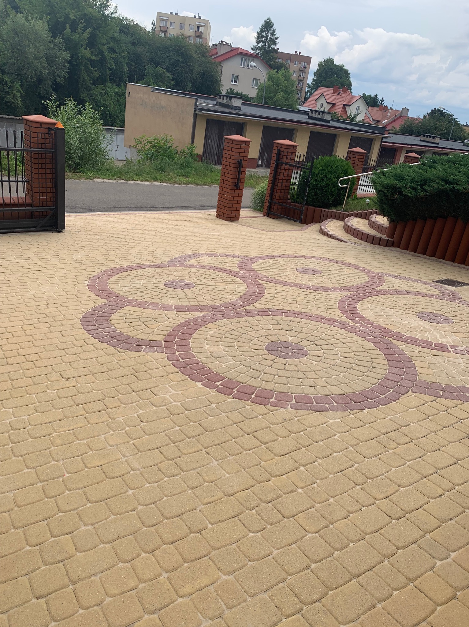 Ułożona kostka brukowa w odcieniach beżu i czerwieni tworząca wzór koncentrycznych okręgów na podjeździe, brama wjazdowa z ceglanymi słupkami w tle.