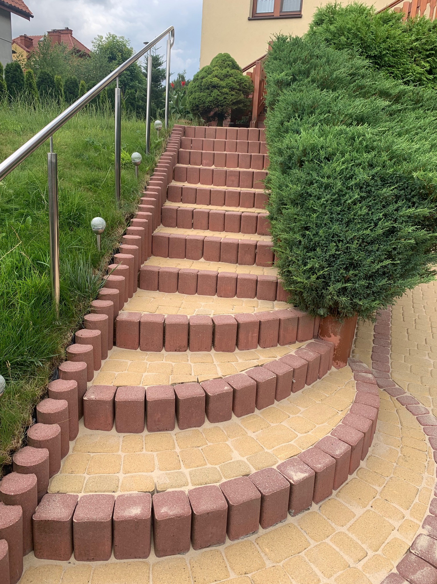 Schody zewnętrzne wyłożone kostką brukową w dwóch kolorach, z metalową barierką i ozdobnymi kulami świetlnymi w trawie, prowadzące do budynku z zielenią po bokach.