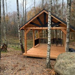 Plan Drewno - Drewniana konstrukcja altany z czarnymi elementami dachu, wkomponowana w leśny krajobraz z brzozami i kamieniami. Widoczne dwa krzesła wewnątrz.