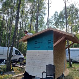 Plan Drewno - Kobieta montuje dach niewielkiej drewnianej konstrukcji w lesie, widoczny częściowo ocieplony bok budynku z membraną Divoroll, obok zaparkowane busy, w dole leży pies.