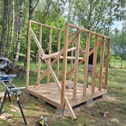 Plan Drewno - Konstrukcja szkieletowa altany w trakcie budowy, widoczne drewniane elementy, podstawa z betonowych bloczków, piła stołowa i ukośnica w tle, teren zalesiony.