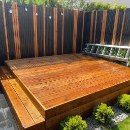 Plan Drewno - Dwupoziomowy taras z drewna o ciepłym odcieniu, otoczony zielenią i ogrodzeniem z pionowych desek w kolorze czarnym i naturalnym, z dekoracyjnymi żarówkami. Przy ogrodzeniu oparta aluminiowa drabina.