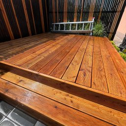 Plan Drewno - Nowo wybudowany taras drewniany z widocznymi schodkami, zabezpieczony lakierem, z nowoczesnym ogrodzeniem z pionowych desek w kolorach brązu i czerni oraz aluminiową drabiną opartą o ogrodzenie.