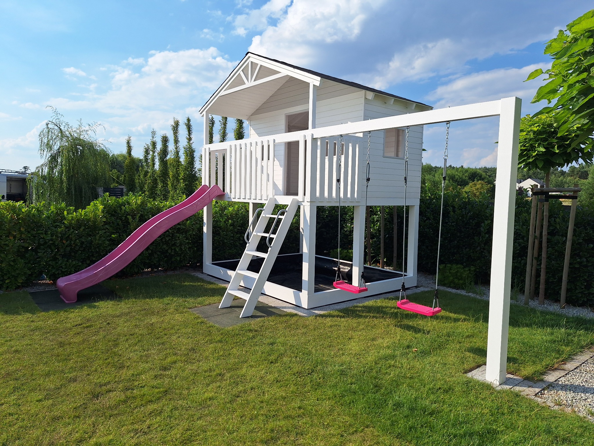 Biały domek na drzewie z różową zjeżdżalnią i huśtawkami na tle zielonego ogrodu i błękitnego nieba z chmurami.