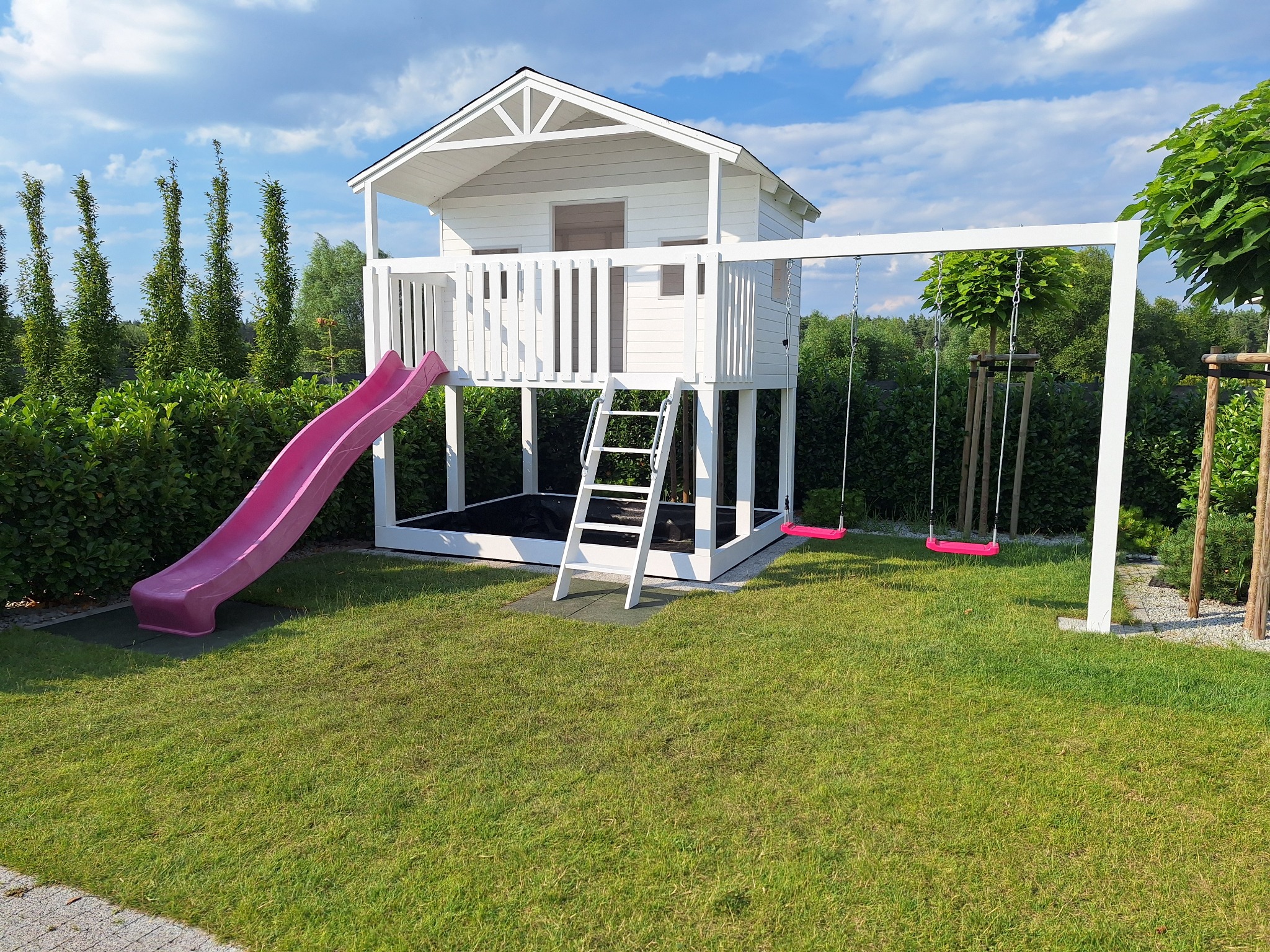 Biały domek dla dzieci z różową zjeżdżalnią i huśtawką na tle zielonego trawnika i drzew