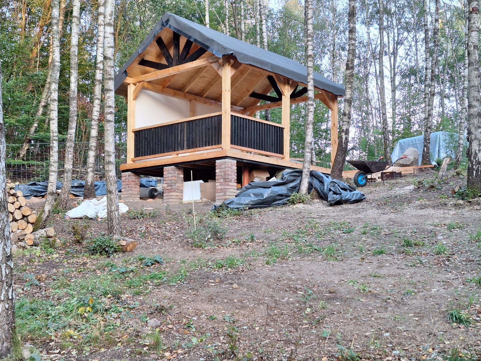Drewniana altana z czarną balustradą, w trakcie budowy, posadowiona na ceglanych fundamentach, otoczona brzozowym lasem, z widocznymi materiałami budowlanymi i narzędziami.