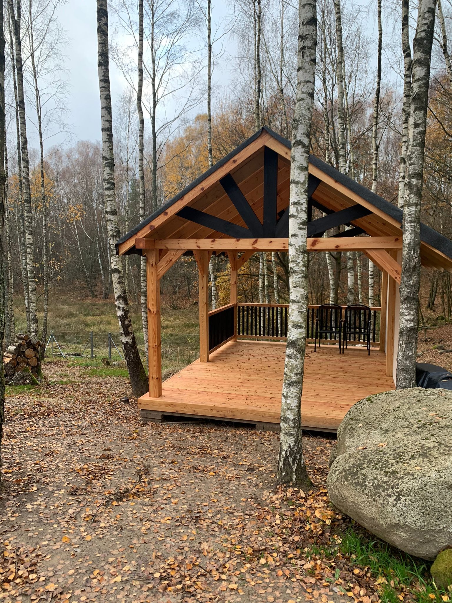 Drewniana konstrukcja altany z czarnymi elementami dachu, wkomponowana w leśny krajobraz z brzozami i kamieniami. Widoczne dwa krzesła wewnątrz.