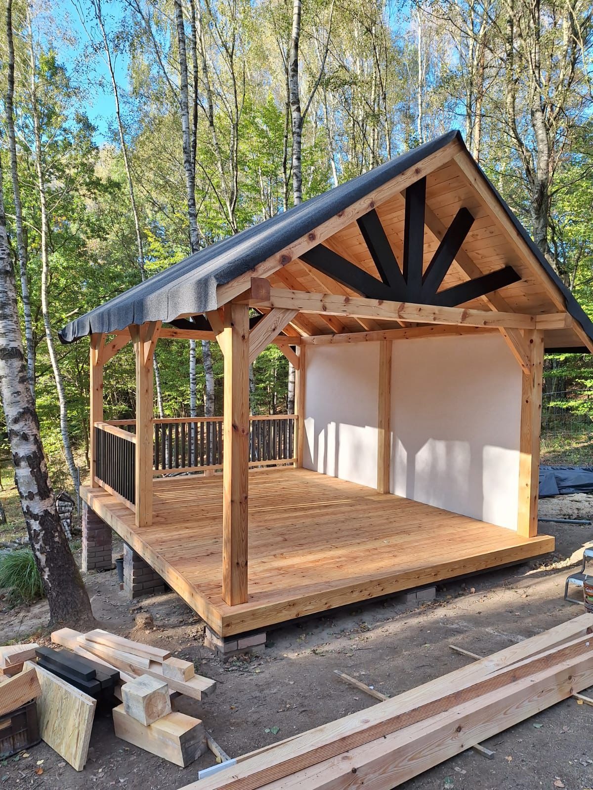 Nowoczesna altana z drewna, z czarnymi elementami dekoracyjnymi w konstrukcji dachu, częściowo obudowana, z drewnianą podłogą, otoczona drzewami.