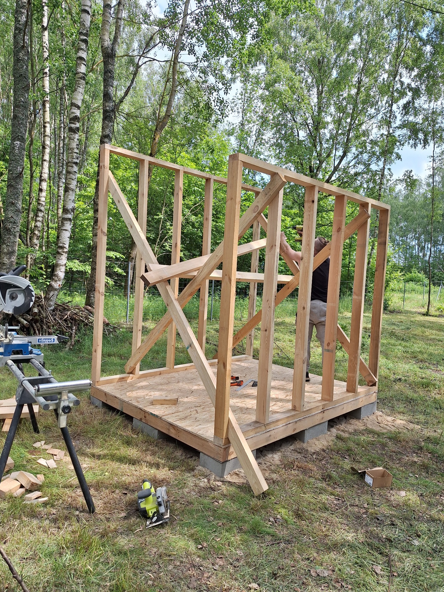 Konstrukcja szkieletowa altany w trakcie budowy, widoczne drewniane elementy, podstawa z betonowych bloczków, piła stołowa i ukośnica w tle, teren zalesiony.