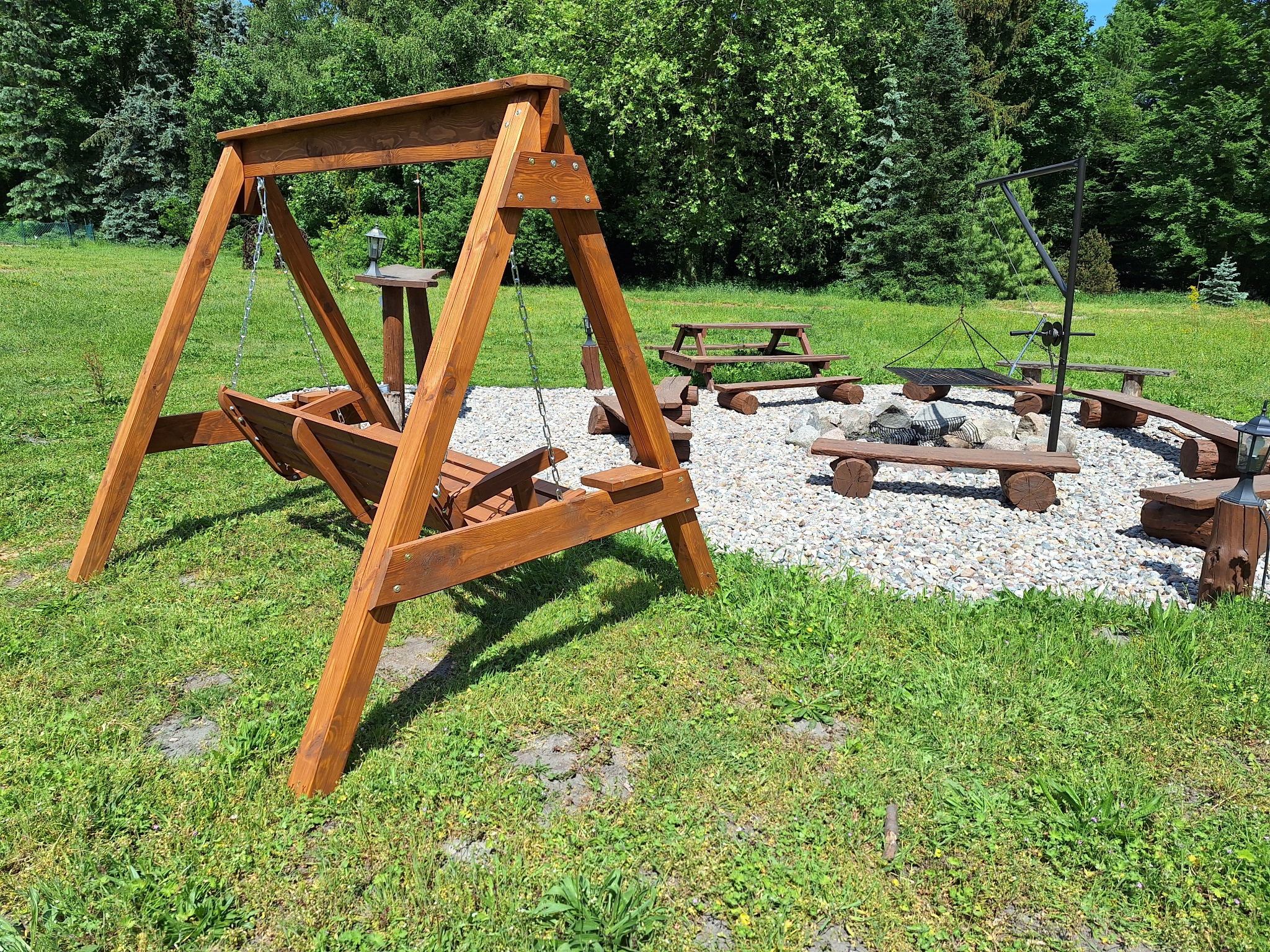 Drewniana huśtawka ogrodowa z ławeczką, w tle miejsce na ognisko z siedziskami z bali i stoliki piknikowe na trawniku.