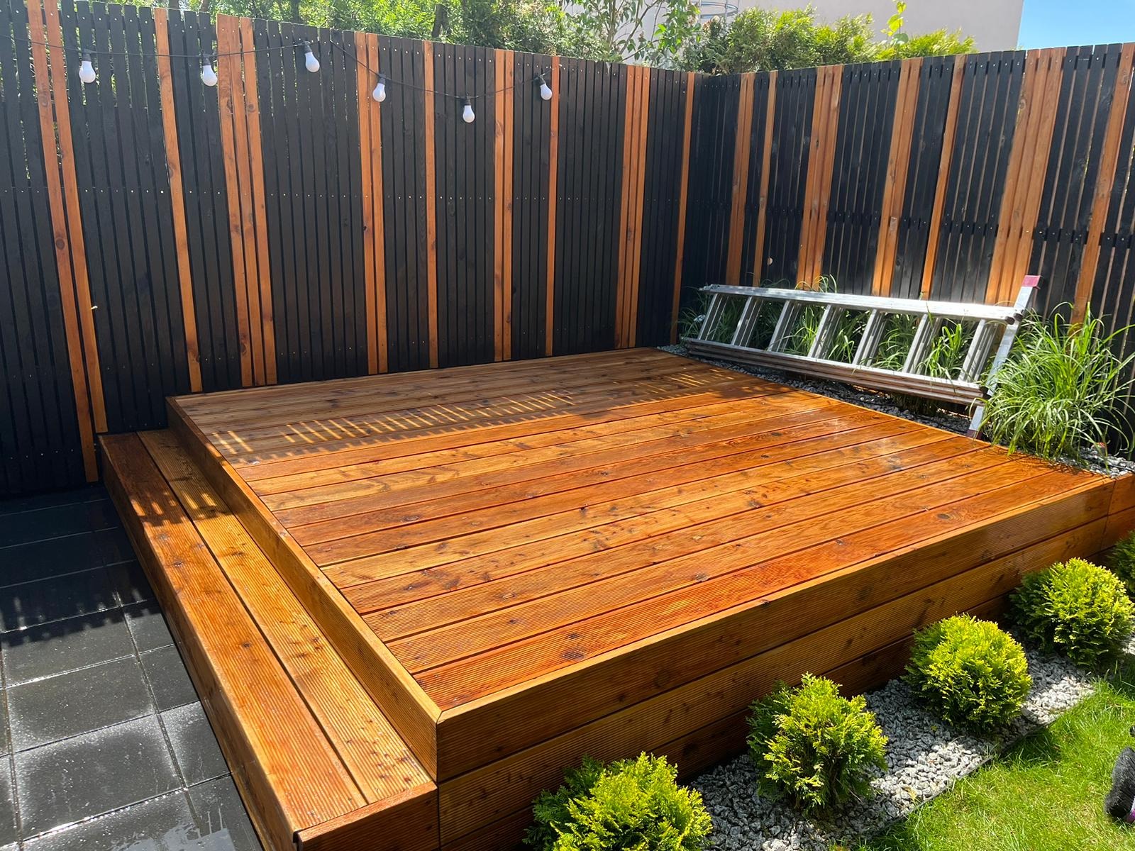 Dwupoziomowy taras z drewna o ciepłym odcieniu, otoczony zielenią i ogrodzeniem z pionowych desek w kolorze czarnym i naturalnym, z dekoracyjnymi żarówkami. Przy ogrodzeniu oparta aluminiowa drabina.