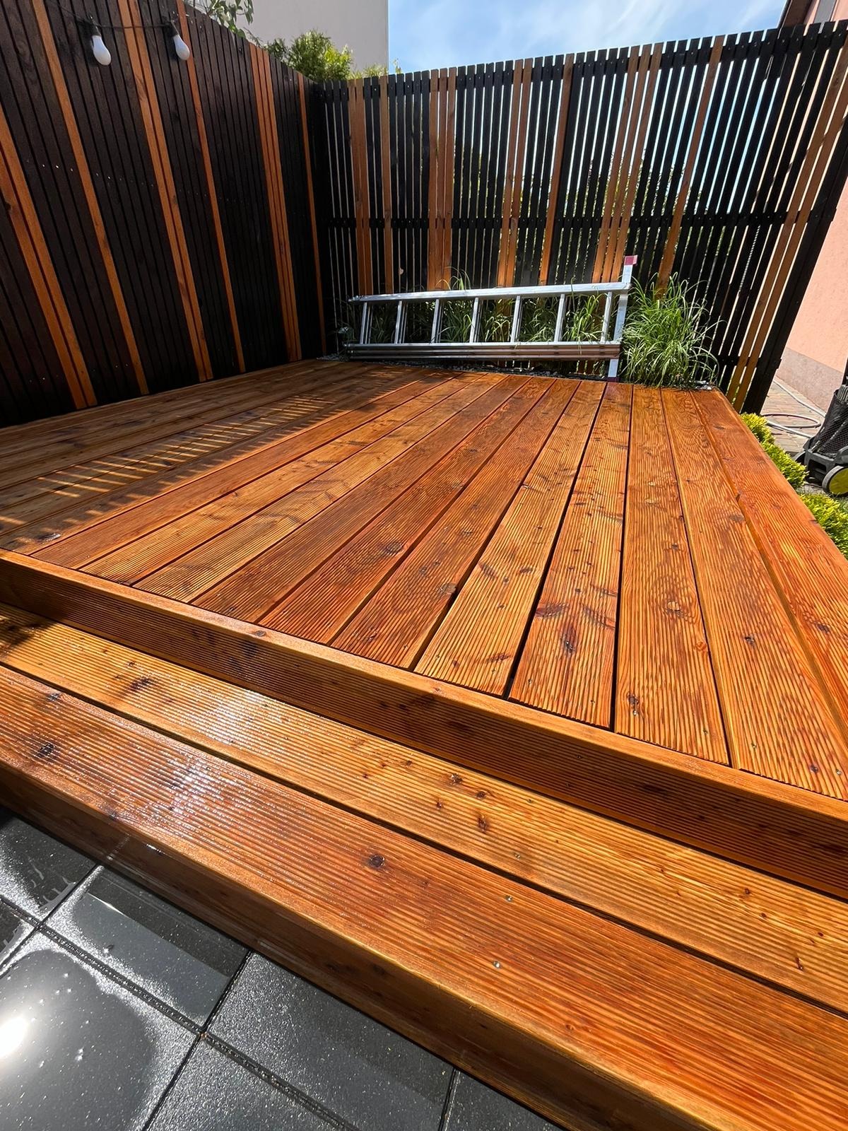 Nowo wybudowany taras drewniany z widocznymi schodkami, zabezpieczony lakierem, z nowoczesnym ogrodzeniem z pionowych desek w kolorach brązu i czerni oraz aluminiową drabiną opartą o ogrodzenie.