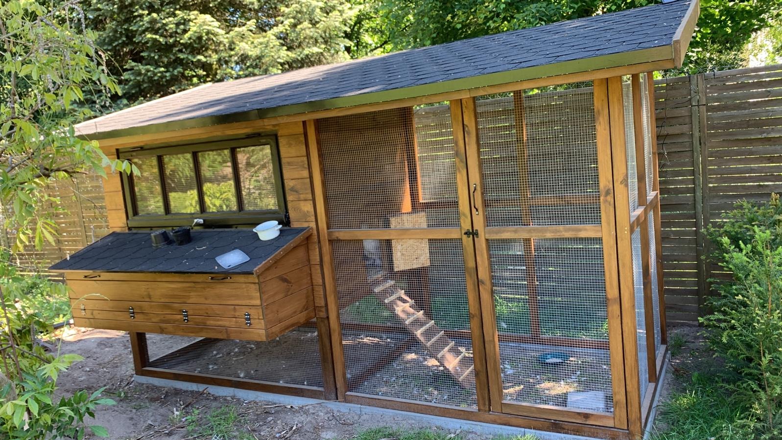 Drewniany kurnik z zadaszeniem i wybiegiem z siatki, umieszczony w ogrodzie, z widocznymi elementami wyposażenia wewnątrz.