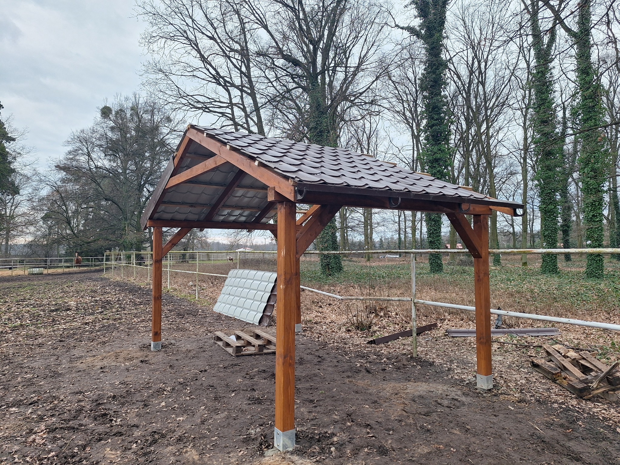 Drewniana konstrukcja z zadaszeniem z brązowej dachówki, prawdopodobnie w trakcie budowy, na tle drzew i pastwiska.