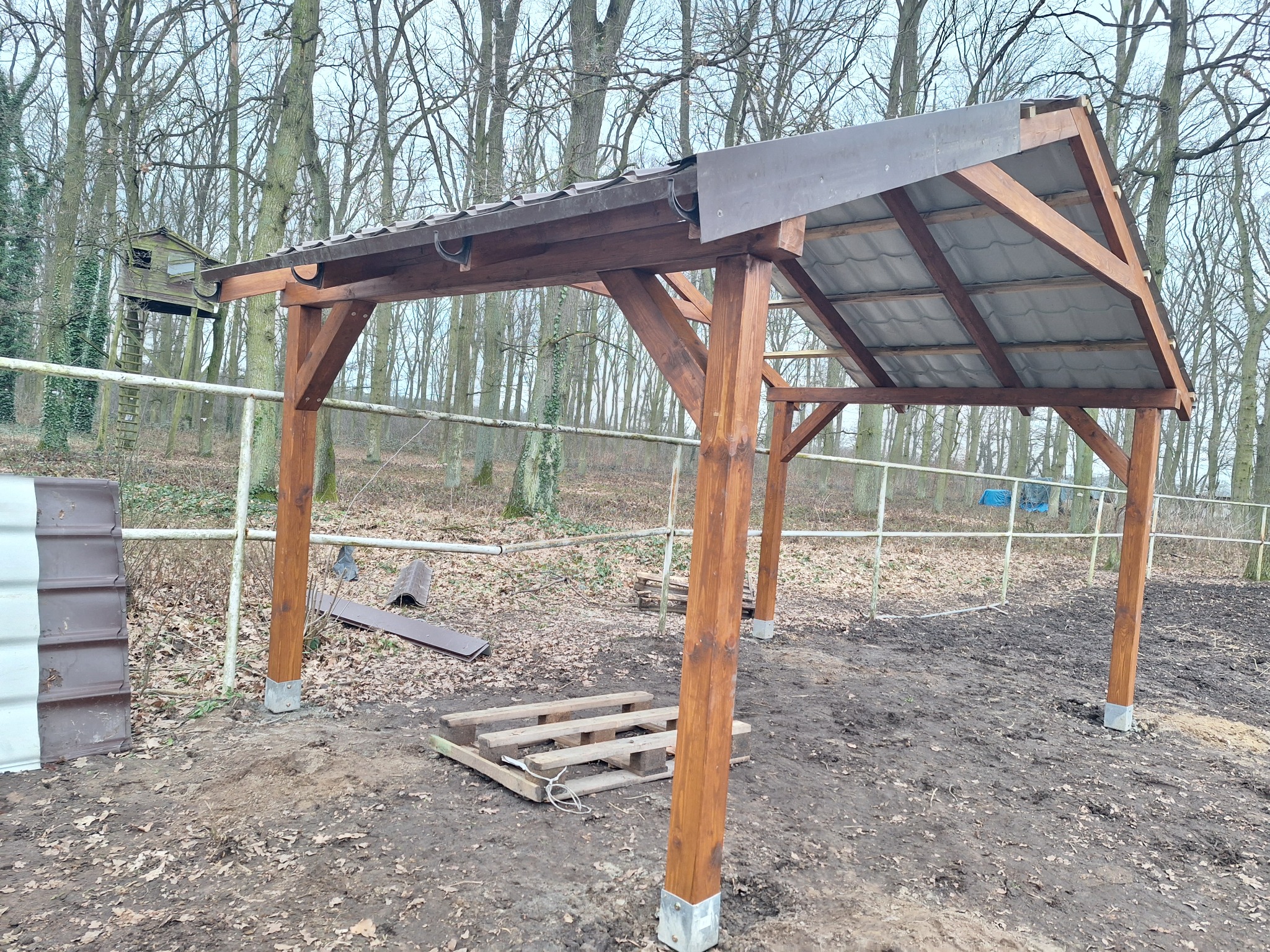 Drewniana konstrukcja z zadaszeniem w trakcie budowy, widoczne palety i elementy konstrukcyjne, tło z drzewami.