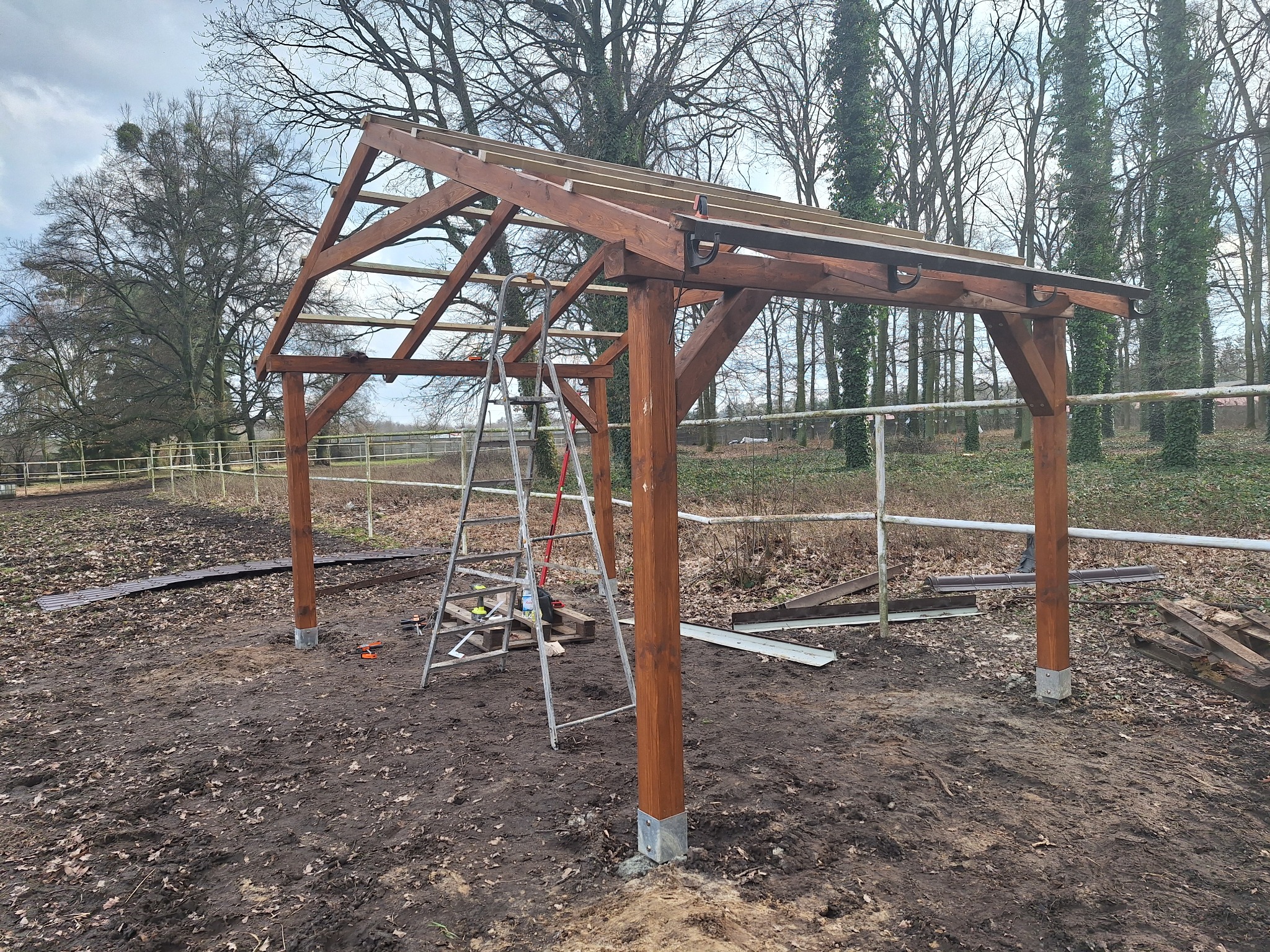 Konstrukcja drewniana w trakcie budowy, widoczny szkielet z brązowych belek i słupów, drabina oparta o konstrukcję, narzędzia leżące na ziemi, tło stanowią drzewa bez liści i pochmurne niebo.